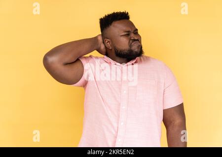 Portrait of worried anxious bearded man wearing pink shirt touching neck, feeling acute pain moving and turning head, suffering spine problems. Indoor studio shot isolated on yellow background. Stock Photo