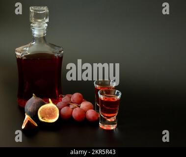 Fruit liqueur on a black background, a bottle and two glasses of homemade alcohol, next to pieces of ripe figs and a bunch of dark grapes. Close-up. Stock Photo