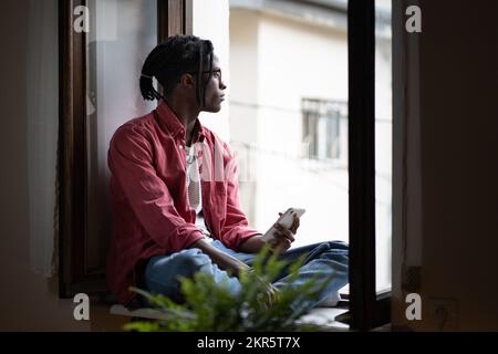 African student guy sitting on windowsill holding smartphone, missing friends while studying abroad Stock Photo