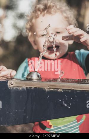 Young blond boy with curly blonde hair watches with amazement of forming and falling water droplets trickle from the facet of a drinking fountain. Cur Stock Photo