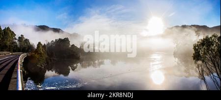 Scenic panorama landscape of early morning mist on Lake Rosebery, in Tullah Tasmania, Australia with the sun bursting above the mountain peaks and ref Stock Photo