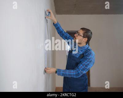 Side view of serious man using measuring tape in room with underfloor heating pipes. Designer in shirt taking measurements while working on renovation Stock Photo