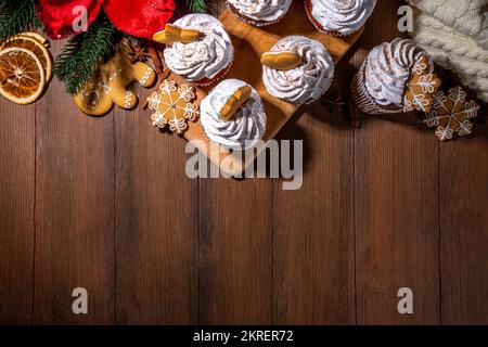 Christmas gingerbread cupcakes. Sweet chocolate and white muffin cupcakes with whipped cream, winter spices and gingerbread cookies decor, on wooden c Stock Photo