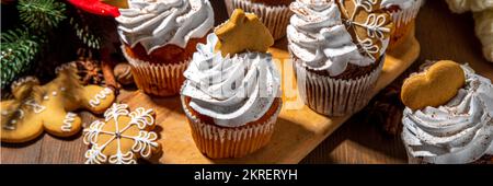 Christmas gingerbread cupcakes. Sweet chocolate and white muffin cupcakes with whipped cream, winter spices and gingerbread cookies decor, on wooden c Stock Photo
