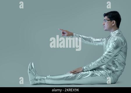 side view of a man wearing silver dress sitting and pointing with his right arm on a grey backdrop, future technology concept. Stock Photo