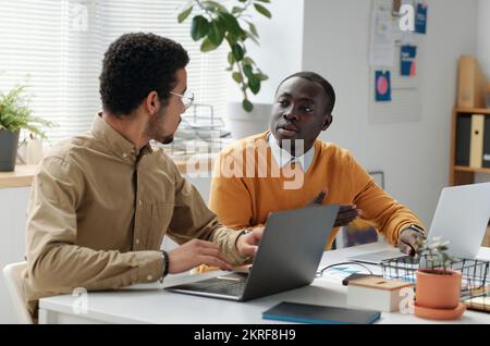 Two young businessmen sitting at table and discussing online project together using their computers Stock Photo