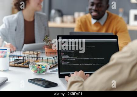 Close-up of young developer sitting at table in front of laptop and working with codes at meeting with colleagues in background Stock Photo