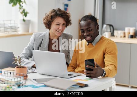 Young colleagues using smartphone for online video call while working at table together with laptop at office Stock Photo