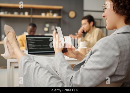 Rear view of young businesswoman typing a message on her mobile phone and smiling while resting at workplace at office Stock Photo