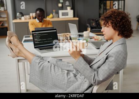 Young businesswoman sitting at her workplace with laptop and using smartphone during break at office Stock Photo