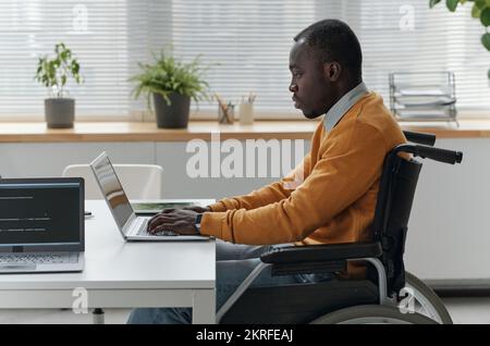 African American developer with disability sitting on wheelchair and working on laptop at his workplace at office Stock Photo