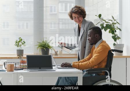 Smiling businesswoman using tablet pc and consulting with programmer with disability while he working on laptop Stock Photo