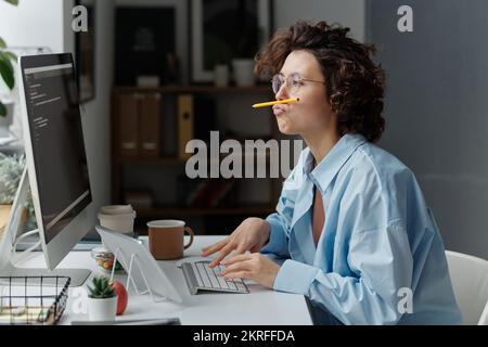 Young businesswoman fooling around while sitting at her workplace in front of monitor and typing codes on computer Stock Photo