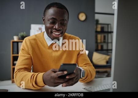 African American businessman typing message on smartphone and smiling while sitting at his workplace at office Stock Photo