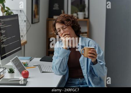 Young tired businesswoman yawning and drinking coffee while sitting at her workplace at office Stock Photo