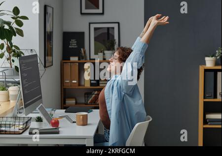 Young businesswoman doing stretching exercises while sitting at her workplace in front of computer monitor Stock Photo