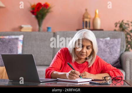 Old woman writing on notepad while using laptop at home Stock Photo