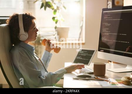 Female programmer in headphones sitting at her workplace in front of computer monitor and doing exercise with dumbbells Stock Photo