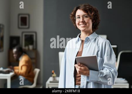 Portrait of young office worker in eyeglasses with tablet pc smiling at camera standing at office Stock Photo