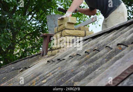 Chimney repair with laying new bricks. Building contractor is bricklaying new chimney pipe on old house with asbestos roof. Stock Photo