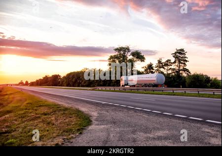 A truck with a semi-trailer tanker transports dangerous goods against the backdrop of a sunny sunset and the sky in the clouds. Transportation of flui Stock Photo