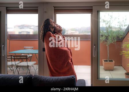 Couple of young gay boys have fun together hugging and wrapped in blanket, staying at home in gray autumn day - people diversity representation lifest Stock Photo