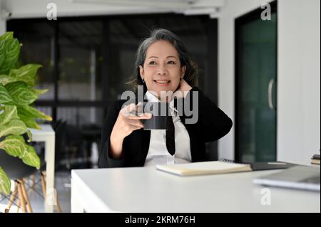 Happy and cheerful Asian senior businesswoman or female boss enjoys sipping coffee and daydreaming while sitting at her desk. Stock Photo