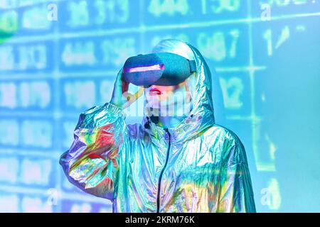 Young blond female in VR headsets standing against wall with glowing neon numbers on unrealistic image admiring Stock Photo