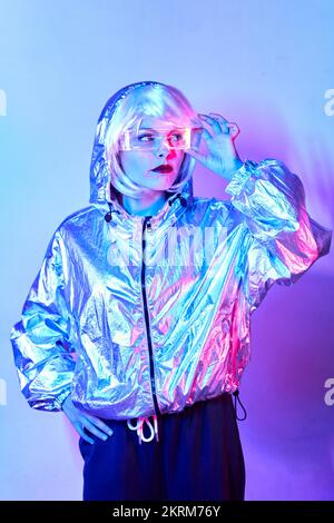 Young female in wig with smart augmented reality goggles with virtual screen displaying icons while standing in studio with glowing neon blue lights Stock Photo