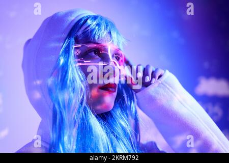 Young female in wig touching smart augmented reality goggles with virtual screen displaying icons while standing in studio with glowing neon blue ligh Stock Photo