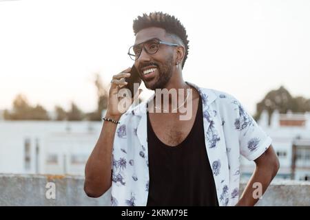 Cheerful African American male in casual outfit and glasses smiling while making a phone call on smartphone outside looking away Stock Photo