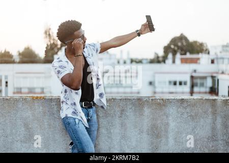 Side view of cheerful African American male in casual wear with wristwatch bracelet and necklace standing outside while taking selfie on smartphone Stock Photo