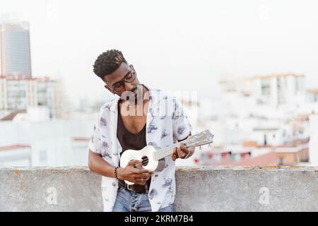 Young African American male musician in casual apparel standing against concrete border while playing song on ukulele Stock Photo