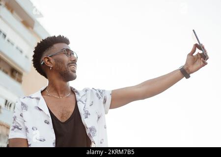 From below of cheerful African American male in casual wear with wristwatch bracelet and necklace standing outside while taking selfie on smartphone Stock Photo