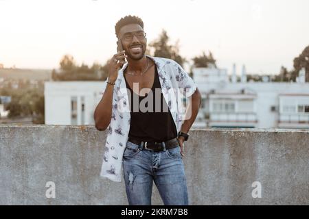 Cheerful African American male in casual outfit and glasses smiling while making a phone call on smartphone outside looking away Stock Photo