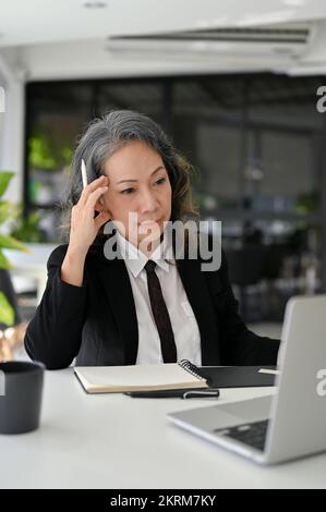 Portrait, Thoughtful Asian senior businesswoman or female executive manager concentrating on her business project, using laptop computer, working in h Stock Photo
