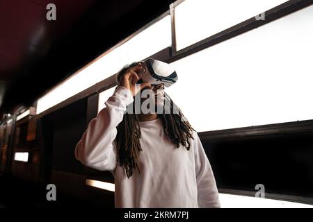Young astonished African American male touching VR goggles experiencing virtual reality in modern room with glowing lamps Stock Photo