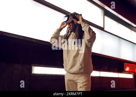 Young astonished African American male touching VR goggles experiencing virtual reality in modern room with glowing lamps Stock Photo