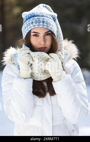 Thoughtful lady in winter clothes looking at camera and touching long tip of cap while standing in snowy forest in sunlight Stock Photo