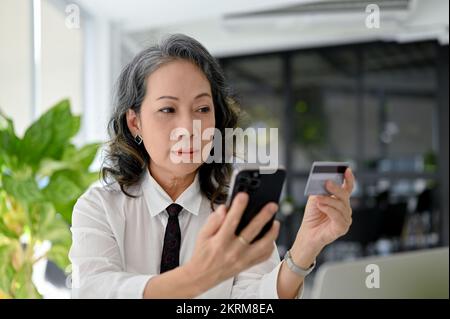 A beautiful Aged Asian businesswoman holding her smartphone and a credit card, using mobile banking application. Stock Photo