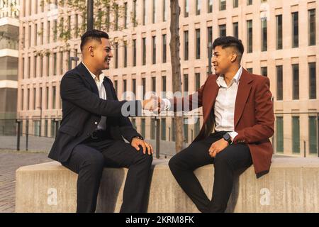 Side view of ethnic men in elegant suits shaking hands while greeting each other sitting on concrete bench Stock Photo