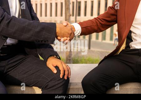 Side view of cropped unrecognizable ethnic men in elegant suits shaking hands while greeting each other sitting on concrete bench Stock Photo