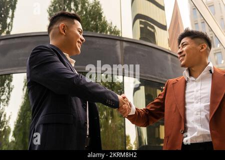 Side view low angle of Hispanic men in formal clothes standing near modern building reflecting city street while greeting each other Stock Photo