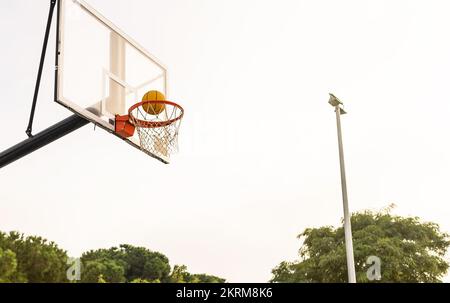 Low angle of orange ball falling into basketball ring with net during game on court against gray sky Stock Photo