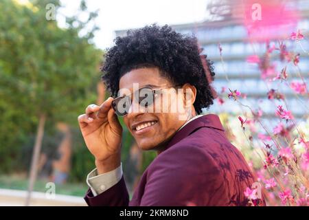 Self assured cheerful young African American male entrepreneur with curly hair in elegant suit and tie and sunglasses looking at camera on city street Stock Photo
