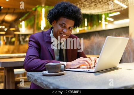 Positive young African American male freelancer with curly hair in stylish suit smiling brightly and looking at camera while working remotely on lapto Stock Photo