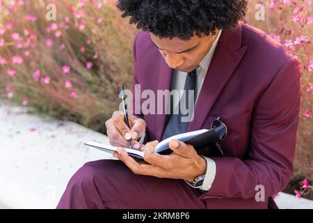Focused ethnic male manager with Afro hair in classy suit taking notes in planner while sitting on bench in park Stock Photo