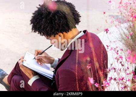 From above side view of focused ethnic male manager with Afro hair in classy suit taking notes in planner while sitting on bench in park Stock Photo