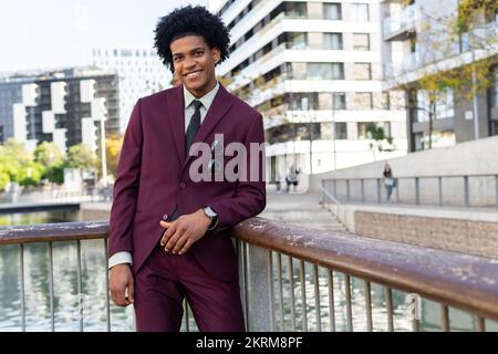 Confident young elegant African American male employee with curly dark hair in classy suit and tie smiling and looking at camera while standing on foo Stock Photo