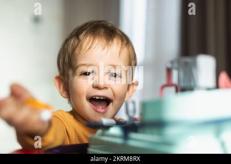 Cheerful adorable little blond haired boy in orange sweater sitting at table and playing with toy while spending time at home Stock Photo